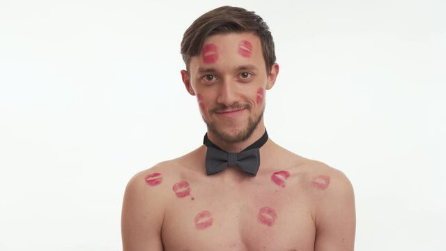 Attractive sexy European half naked brunette man 20s having beard and mustache covered with lipstick prints. Young guy smile, flirt, wink, look up isolated on white wall background. Romantic concept.