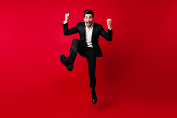 Fototapeta na wymiar Man in elegant suit fooling around in studio. Emotional male model jumping on red background.