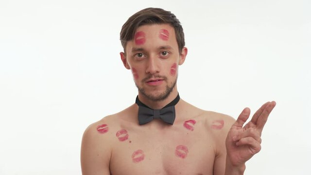 Blow kiss. Close up portrait of handsome young brunette man with beard wearing bow tie shirtless with red lipstick marks on body isolated on white wall background. Sexy male smile, looks at the camera