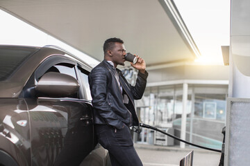 Side view of young handsome African American man, wearing casual black outfit, drinking take away coffee at petrol station, standing leaning on his modern car and refueling it.