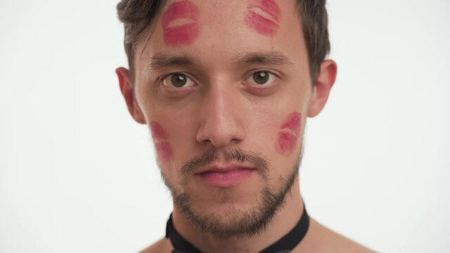 One young Caucasian brunette man model with beard, blow kiss, looks directly at camera on white wall background close up. Cute person kissed by girlfriend with red lipstick kiss marks on the face.
