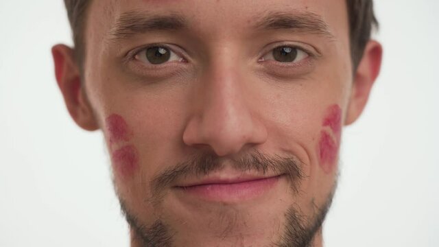 One young Caucasian brunette man model with moustache looks directly at camera isolated on white wall background close up indoor. Handsome guy kissed by girlfriend with red lipstick kiss marks on face