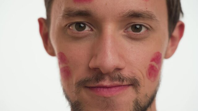 Cute young Casanova boy with beard and moustache, with red lipstick kiss marks on face. Close up portrait of beautiful man stare straight ahead on white background. International Women’s day concept.