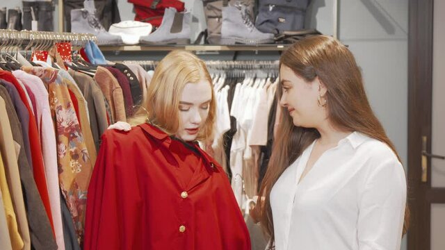 Female friends trying new clothes at fashion store. Young woman trying dresses at clothing boutique, talking to her friend. Shopaholic, spending money concept
