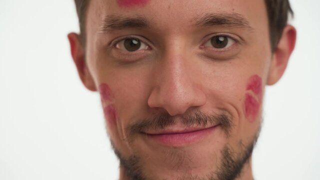 Young beautiful Caucasian bearded man shows kiss spots of red lipstick by girlfriend on the left, right cheeks isolated on white background close up. Portrait of cute guy in kisses. Romantic concept.
