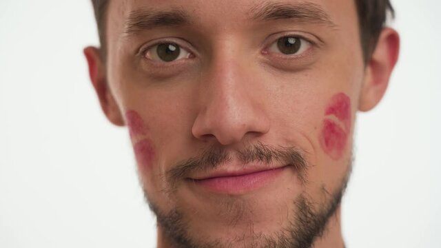 One handsome European brunette man with beard shows red lipstick kiss imprints or marks on left, right cheeks, stare straight isolated on white background close up. International Women’s Day Concept.