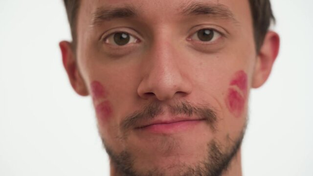 Cute sexy brunette man kissed by girlfriend with red lipstick kiss prints or marks on face looks around on white wall background close up. Handsome bearded guy shy, smile, denies, say not about date.