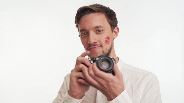 One handsome brunette man with red kiss lips by girl wearing shirt, holding camera in hands, smiling, standing on white wall background close up. Pleased male is photographer. Confident guy portrait