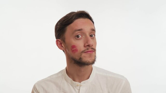 One attractive brunette man with beard wears shirt, looks straight ahead, wipes the kiss mark from his cheek on white background close up. Concept of unfaithfulness on woman. Portrait of one lover guy
