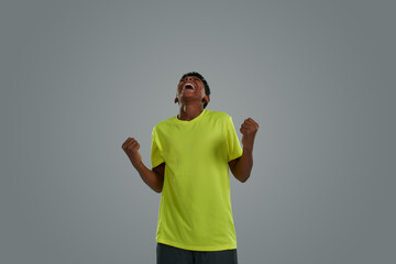 Excited happy teenage african boy wearing sports clothes celebrating success, enjoying victory while standing against grey background