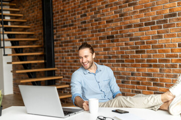 Relaxed millennial young freelancer sitting in the home office with his legs on the desk, completed the project and taking coffee break, holding a cup of coffee and watching funny video on the laptop