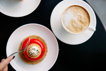 A piece of red glazed mousse cake on a fork. Sponge cake and spongy dessert. Desserts are red. Composition of a sweet snack in a cafe. Aromatic cappuccino coffee