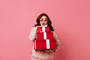 Fototapeta premium Amazed ginger lady holding new year presents on pink background. Studio photo of good-looking girl with gifts.