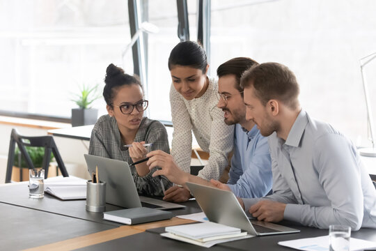 Mixed raced millennial employees talking at shared desk with laptops. Mentor showing and explaining report to interns or students. Marketing team meeting and discussing online video presentation