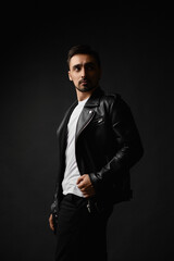 Young handsome man with unshaven face in black leather jacket posing in the dark background, isolate. Stylish bearded guy in a leather jacket