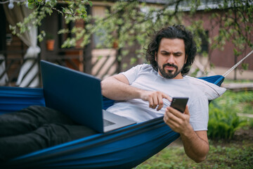 A man works with a laptop and a phone in a hammock in a country house