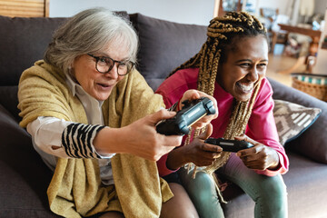 Senior and young woman playing console video games at home during lockdown isolation - Focus on...