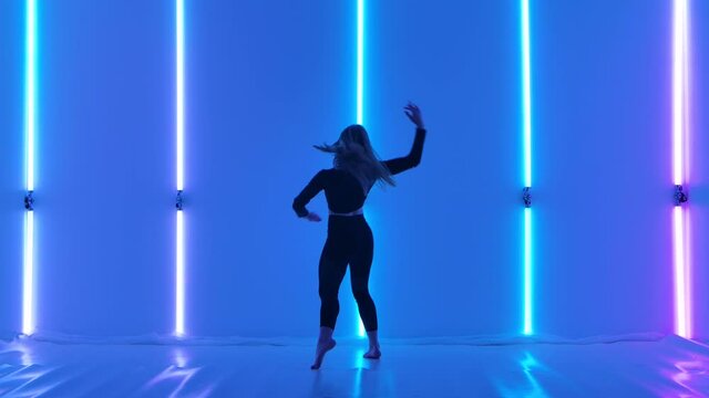 Flexible dancer performs acrobatic elements of modern choreography and jumping against a backdrop of purple neon lights. Sensual woman practices contempt in twilight studio. Slow motion.