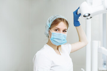 Fototapeta na wymiar Portrait of young female dentist wearing surgical mask examines a patient while holding dental lamp