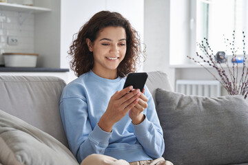 Happy hispanic teen girl holding cell phone using smartphone device at home. Smiling young latin...