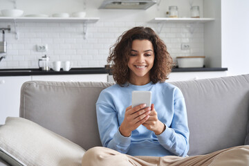 Happy hispanic teen girl looking at smartphone relaxing on couch at home, enjoying using online mobile apps technology, playing games on cell phone, checking messages or social media posts. - obrazy, fototapety, plakaty