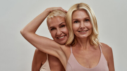 Studio portrait of two happy blonde women smiling at camera, standing together, posing isolated over gray background