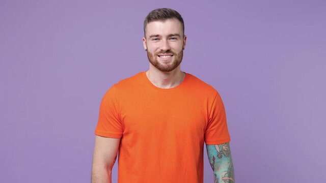 Smiling handsome bearded tattooed young man 20s years old in orange basic casual t-shirt isolated on purple violet color background in studio. People sincere emotions lifestyle concept. Looking camera