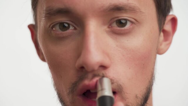 Closeup portrait of handsome bearded young sexy brunette man with moustache getting makeup isolated on white background. Handsome guy looking camera and using lipstick for paints lips in a pink color.
