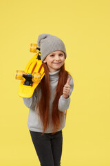 funny little child girl with yellow skateboard is showing thumbs up over yellow background