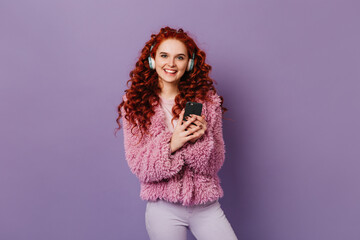 Fototapeta premium Blue-eyed sweet girl looks at camera on purple background with smile. Curly redhead woman in pink jacket posing in headphones with phone in her hands