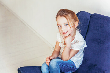 Sweet little girl in jeans and white T-shirt sitting on modern cozy blue chair relaxing in white bright living room at home indoors. Childhood schoolchildren youth relax concept.