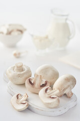 Fototapeta na wymiar Champignons on a white wooden board in the foreground. In the background, in blur, a bowl of chopped mushrooms and a sauce bowl.