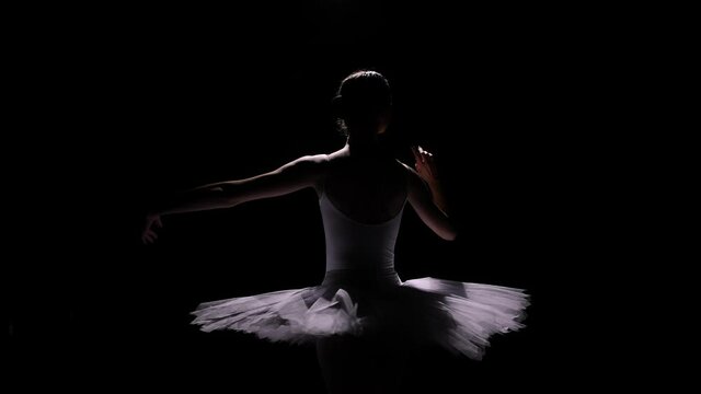 Camera rotates around gentle ballerina in a white tutu performing graceful movements with her hands. Orbital shot of a young dancer soaring against a black studio background. Close up. Slow motion.