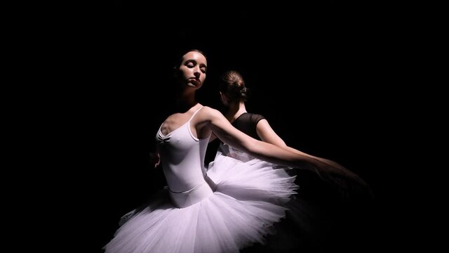 Orbital shot of a pair of charming ballerinas in the form of a black and white swan. Young slender dancers soar on a black studio background with backlight. Silhouettes. Close up. Slow motion.