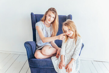 Two happy kids sitting on cozy blue chair relaxing playing in white living room indoors. Little girl playing with teenage girl showing her love care. Sisters having fun at home