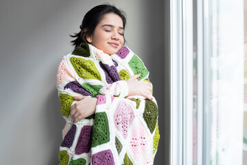 Young woman looking through the window at home wrapped in warm comfy blanket. Self love and self...