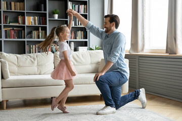 Full length happy young daddy standing on one knee, twisting smiling adorable preschool kid daughter in princess dress, practicing waltz or preparing performance, dancing together in living room.