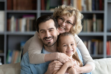 Obraz premium Devoted 60s elderly retired woman in eyeglasses cuddling handsome smiling grown son and small preschool granddaughter, posing together for family photo, enjoying love tender moment in living room.