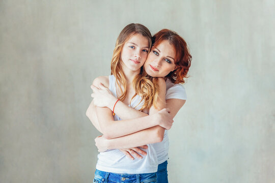 Young Mother Embracing Her Child. Woman And Teenage Girl Relaxing In White Bedroom Near Gray Wall Indoors. Happy Family At Home. Young Mom Playing With Her Daughter