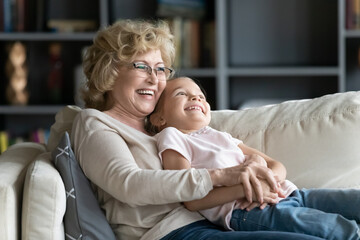 Happy relaxed middle aged senior woman lying on cozy sofa with joyful adorable preschool kid...