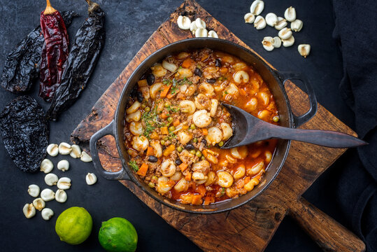 Traditional slow cooked Mexican pozole rojo soup with ground minced beef, hominy maiz and dry ancho paprika offered as top view in cast-iron roasting dish on old rustic wooden board