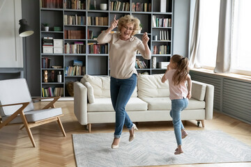 Full length overjoyed mature senior grandmother dancing to disco music with cute little preschool granddaughter, having fun together in living room, involved in energetic domestic activity on weekend.