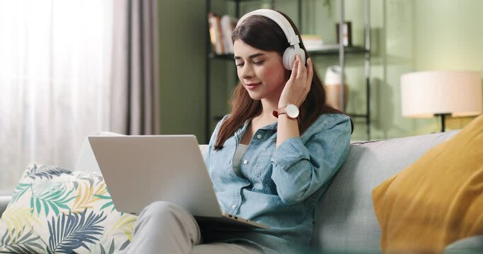 Young beautiful student girl sitting at home on couch typing on laptop enjoying music in white headphones.
