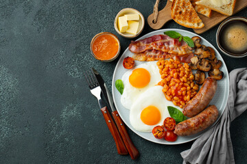 Full English breakfast on a plate with fried eggs, sausages, bacon, beans, toasts and coffee on dark stone background. With copy space. Top view
