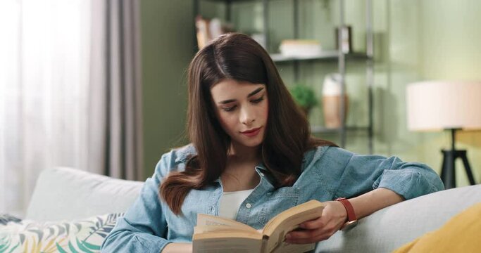Young beautiful caucasian girl spends free time at home sitting on couch and reading interesting book. Concept of self-education.