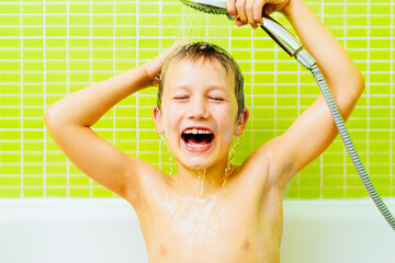 Boy in the bathtub washes his head with water sprayed from the shower head, with funny expression.