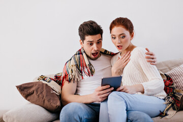 Surprised man and woman posing with tablet. Young couple sitting on cozy sofa with wireless gadget.