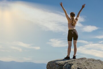 The young woman at the top of the mountain raised her hands up on blue sky background. The woman...