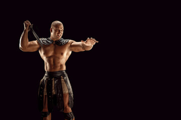 Fototapeta na wymiar Severe barbarian in leather costume with sword. Portrait of balded muscular gladiator. Studio shot. Black background.