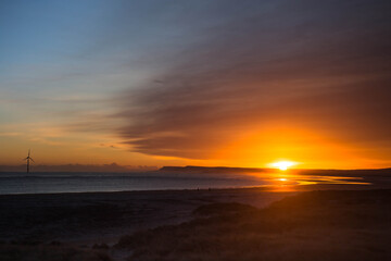 Calm ocean with powerful sunrise at deserted sandy beach with reflections. Wind turbine in background